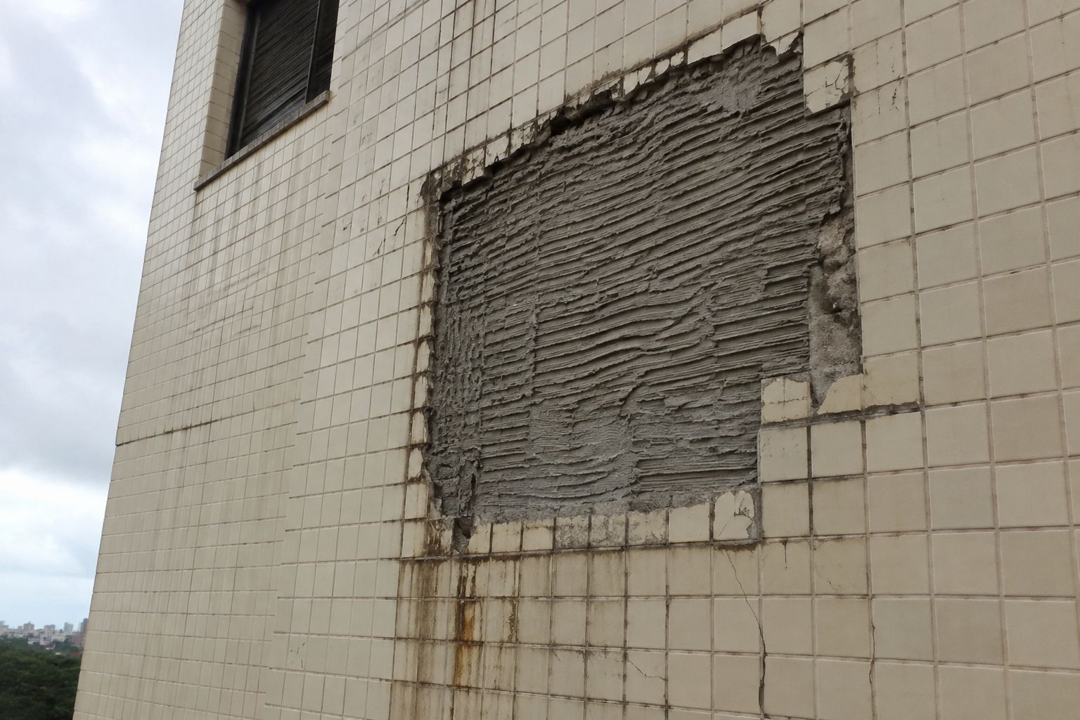 Trecho de fachada com pastilhas cerâmicas claras onde uma área inteira se soltou, deixando à mostra a argamassa colante com os cordões da desempenadeira intactos e rejunte trincado ao redor