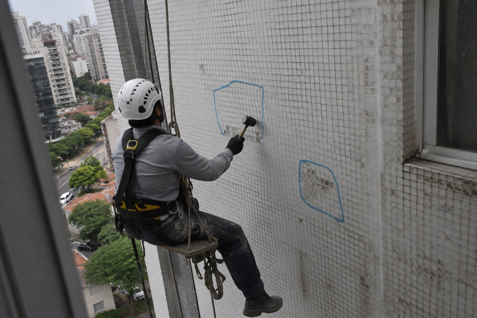 Técnico em acesso por corda, com capacete, cinto paraquedista e cadeirinha, batendo com martelo de borracha nas pastilhas da fachada; áreas com som cavo contornadas a giz azul