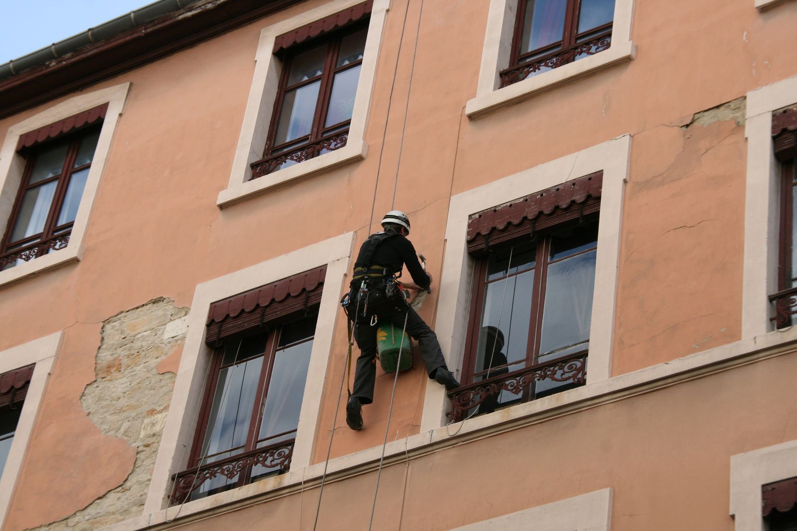 Técnico com capacete e cinto, suspenso por cordas, verifica e remove partes soltas do revestimento na fachada de um edifício